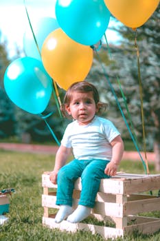 一个可爱的蹒跚学步的孩子坐在阳光明媚的公园里，身旁是一个气球板条箱