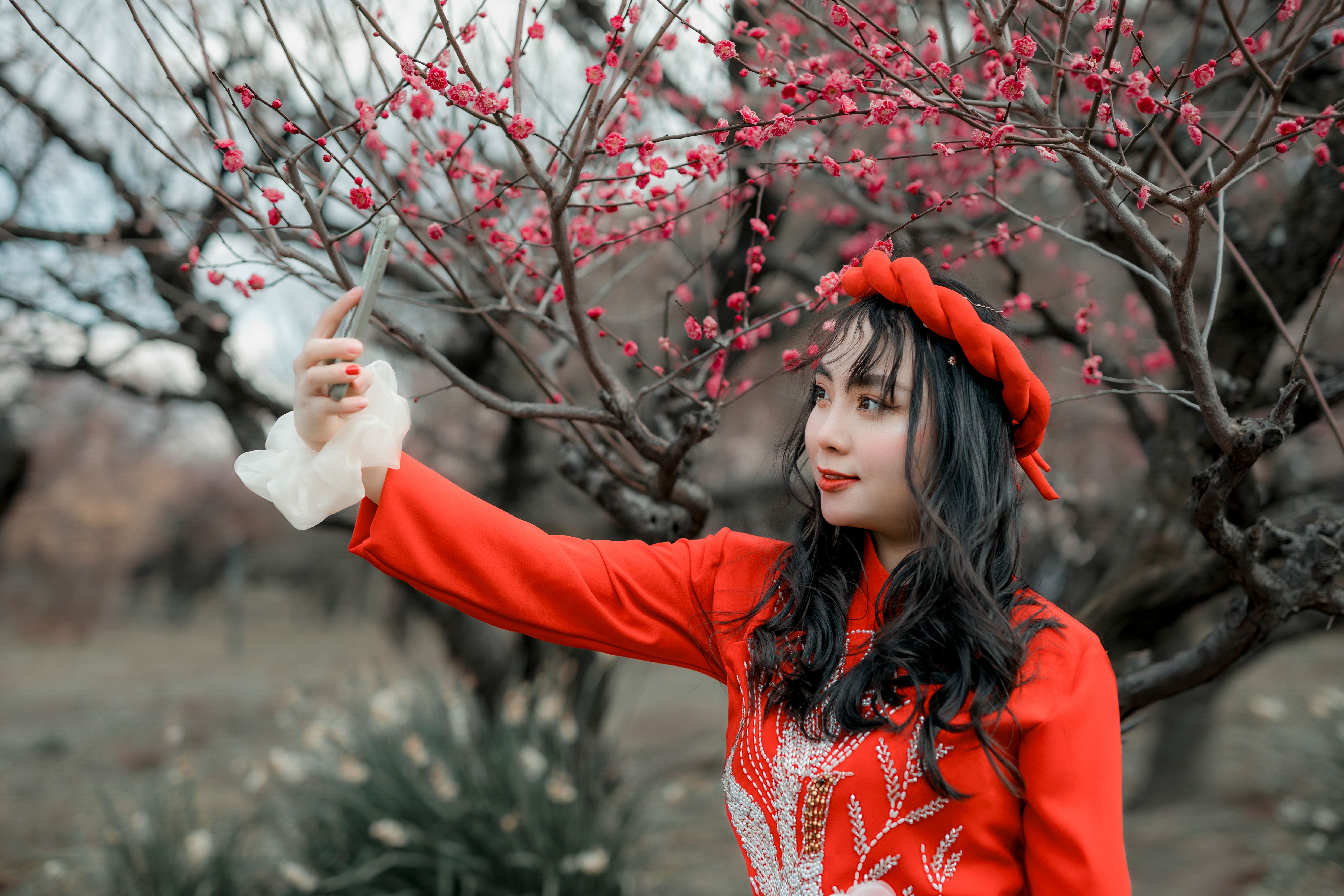 A beautiful Asian woman in red takes a selfie among blossoming pink cherry trees outdoors.