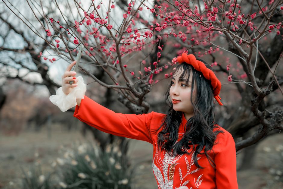 A beautiful Asian woman in red takes a selfie among blossoming pink cherry trees outdoors.