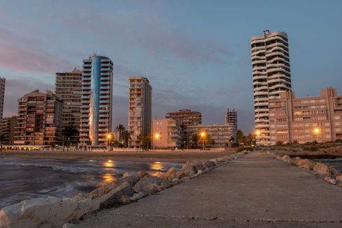 Free Stunning coastal cityscape at dusk with illuminated buildings reflecting on calm waters. Stock Photo