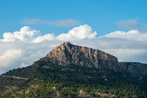 Free A majestic rocky mountain surrounded by lush greenery under a clear blue sky with fluffy clouds. Stock Photo
