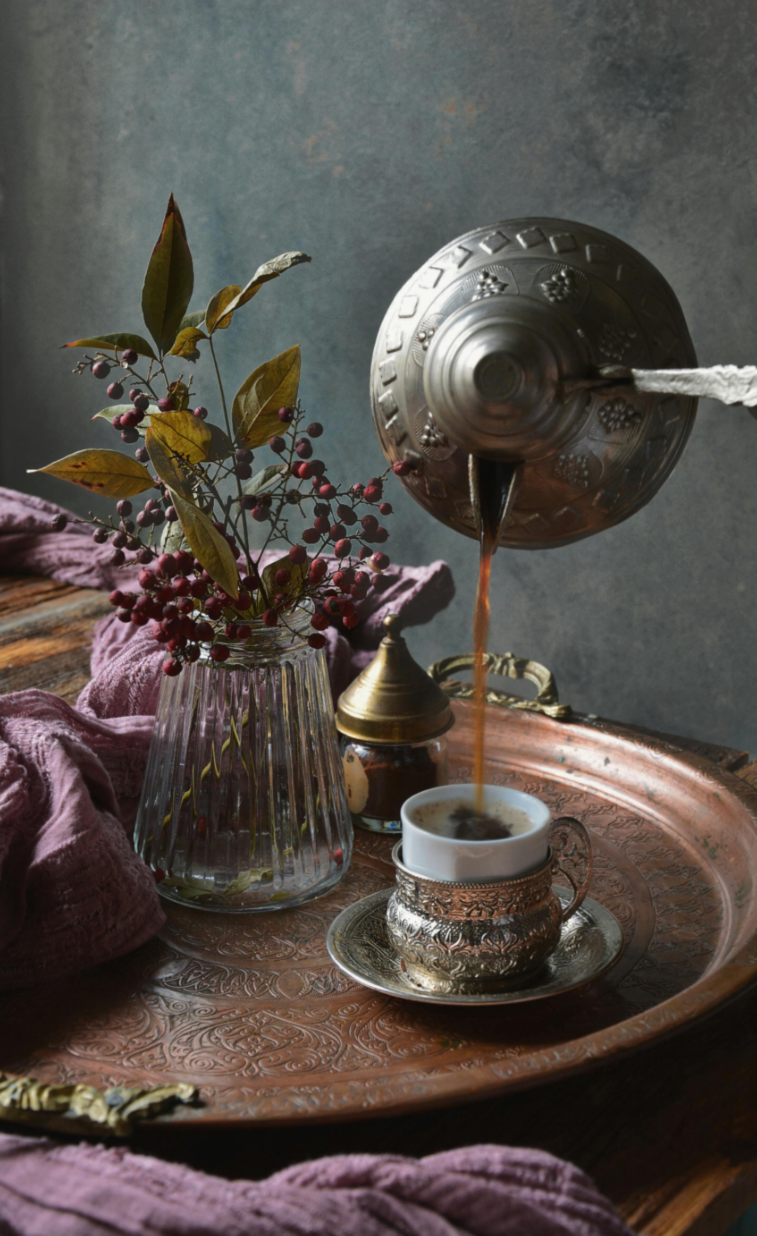 Ornate coffee pot pouring into a decorative cup on a vintage tray with herbs and glass vase.
