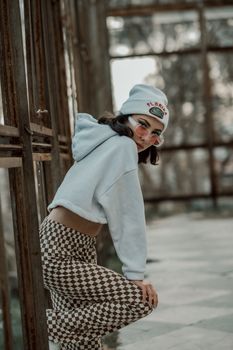 Stylish young woman in a hoodie and beanie posing outdoors with checkered pants.
