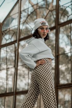 Fashionable woman posing indoors in checkered pants and hoodie under glass ceiling.