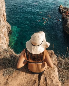 Rear view of a woman in a sun hat sitting on a cliff in Croatia, enjoying a summer day by the Adriatic Sea.