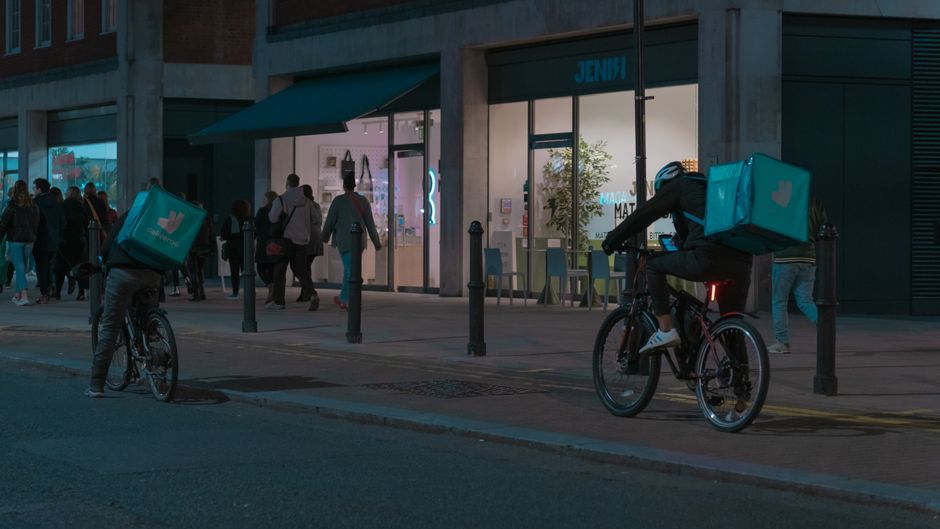 夜の市街地で、自転車の配達作業員が現代的な店舗の近くで歩行者とともにいます。