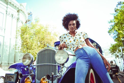 Fashionable young woman posing confidently with a classic blue car on a sunny street.