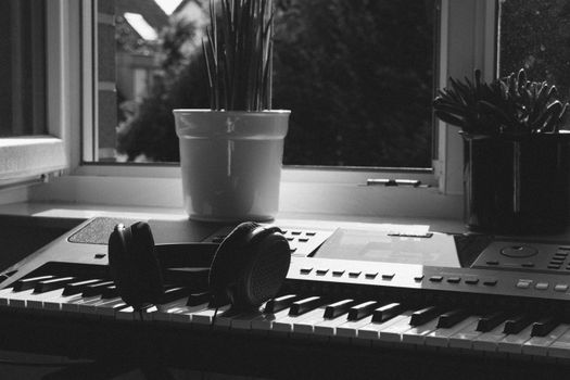 A monochrome photo of a keyboard and headphones near a window with plants, capturing a soothing indoor vibe.