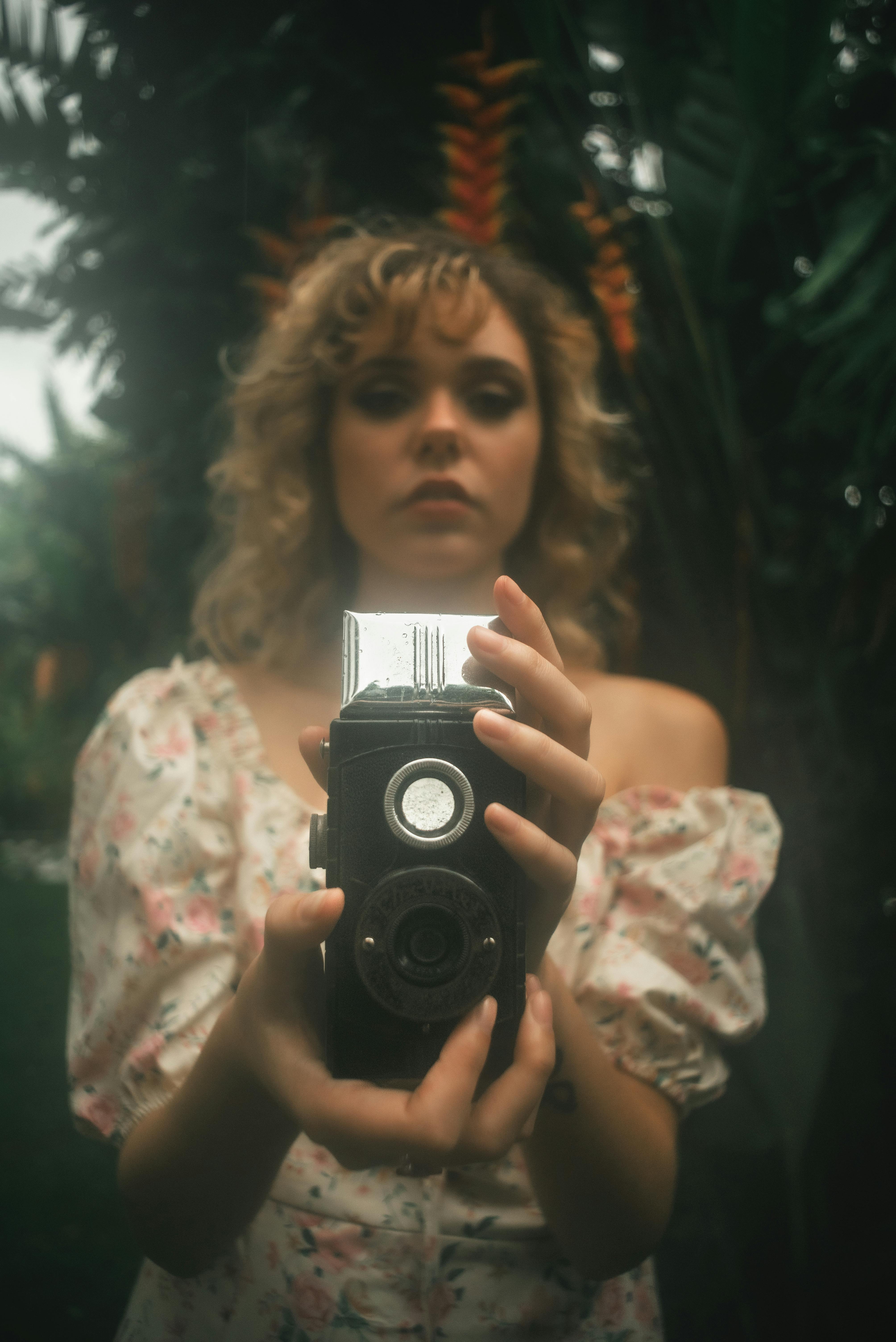 Woman holding a vintage camera with a lush green backdrop, creating a nostalgic and artistic vibe.