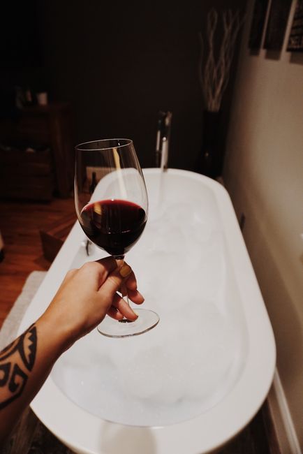 A serene scene of a person holding a glass of red wine next to a bubble-filled bathtub.