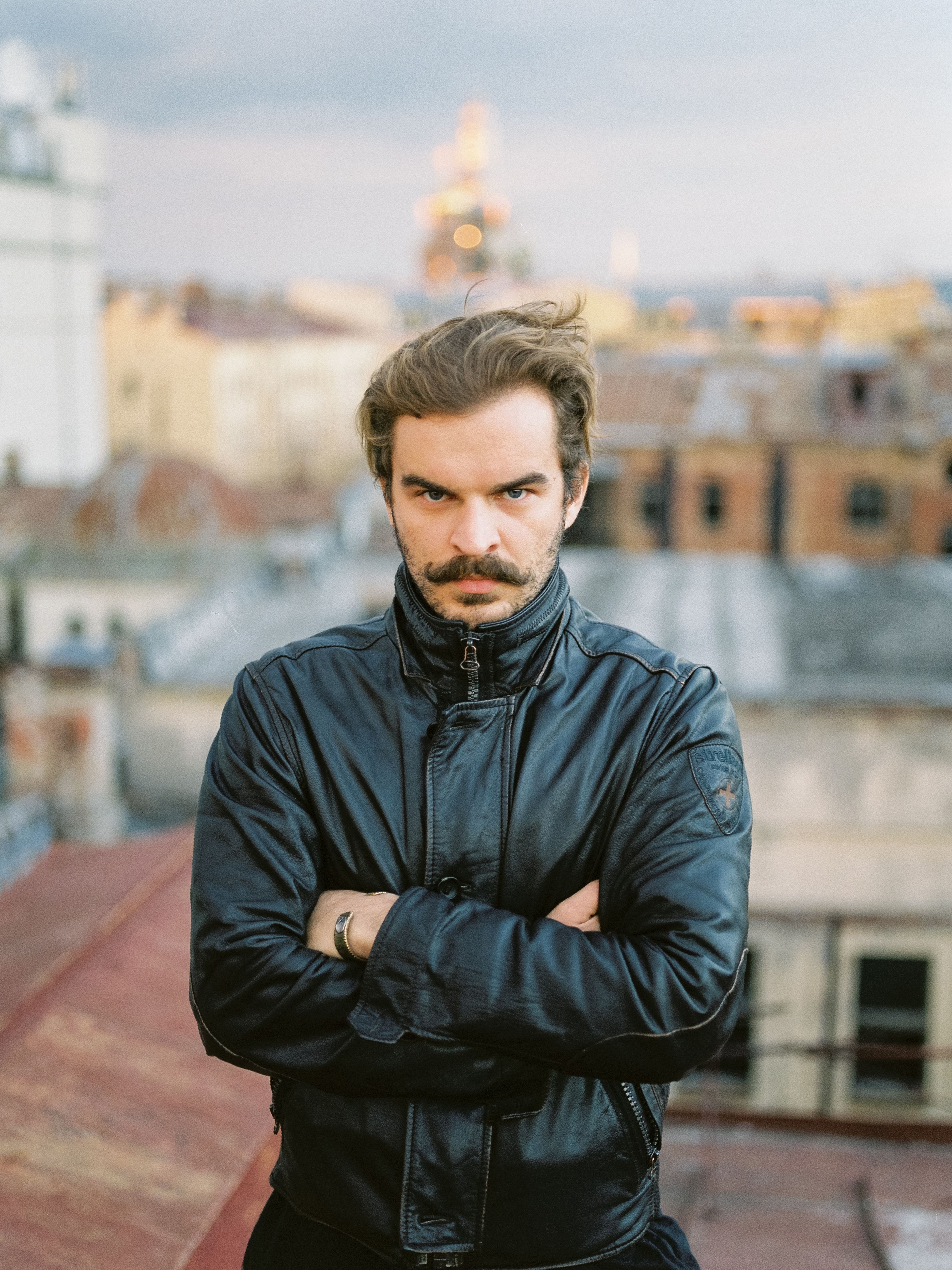 Portrait of a man with a moustache in a leather jacket standing on an urban rooftop with a moody expression.