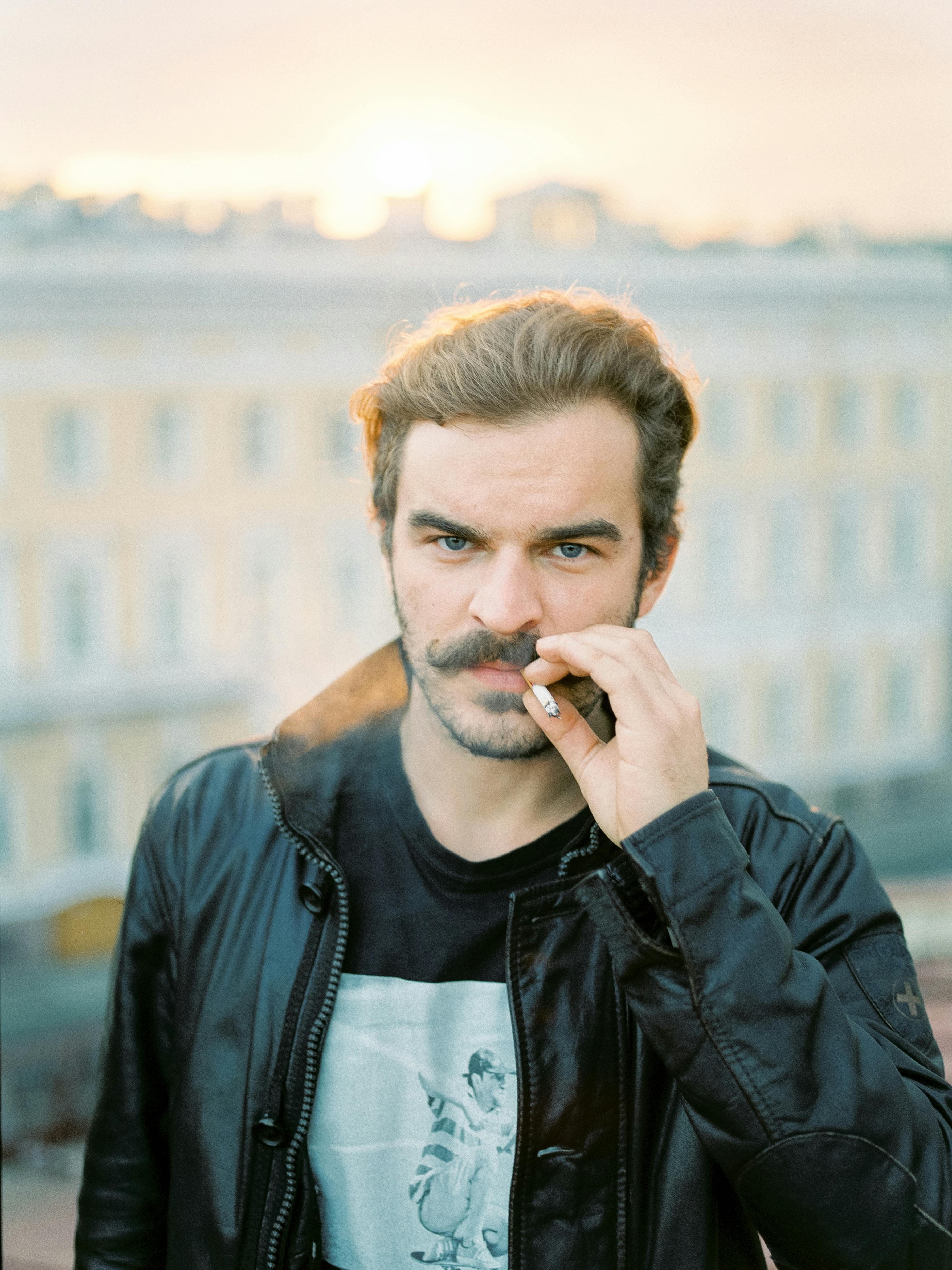 A stylish man smokes a cigarette outdoors, wearing a black leather jacket in an urban setting.