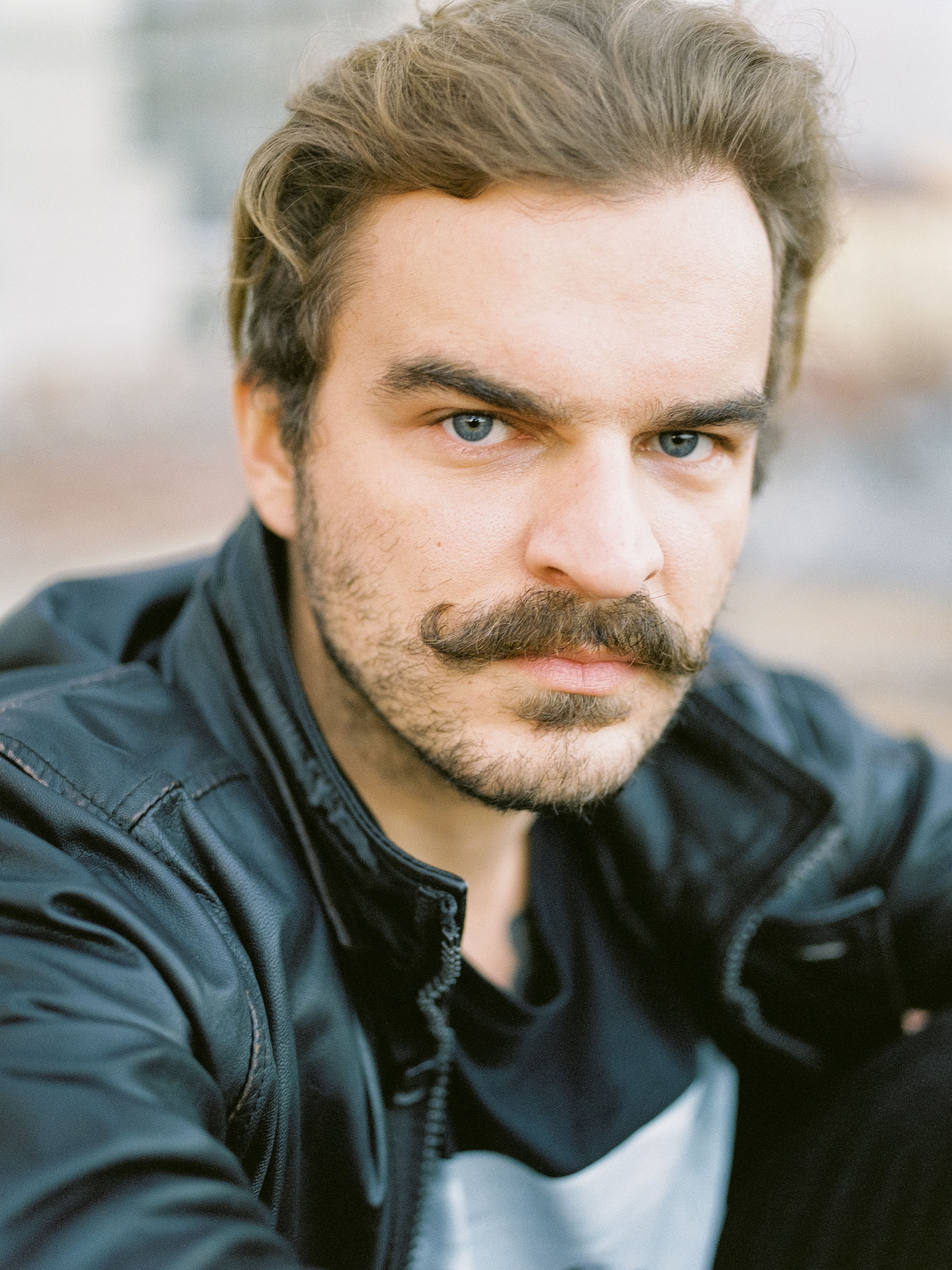 Close-up of a man with a mustache in a leather jacket, looking directly at the camera.