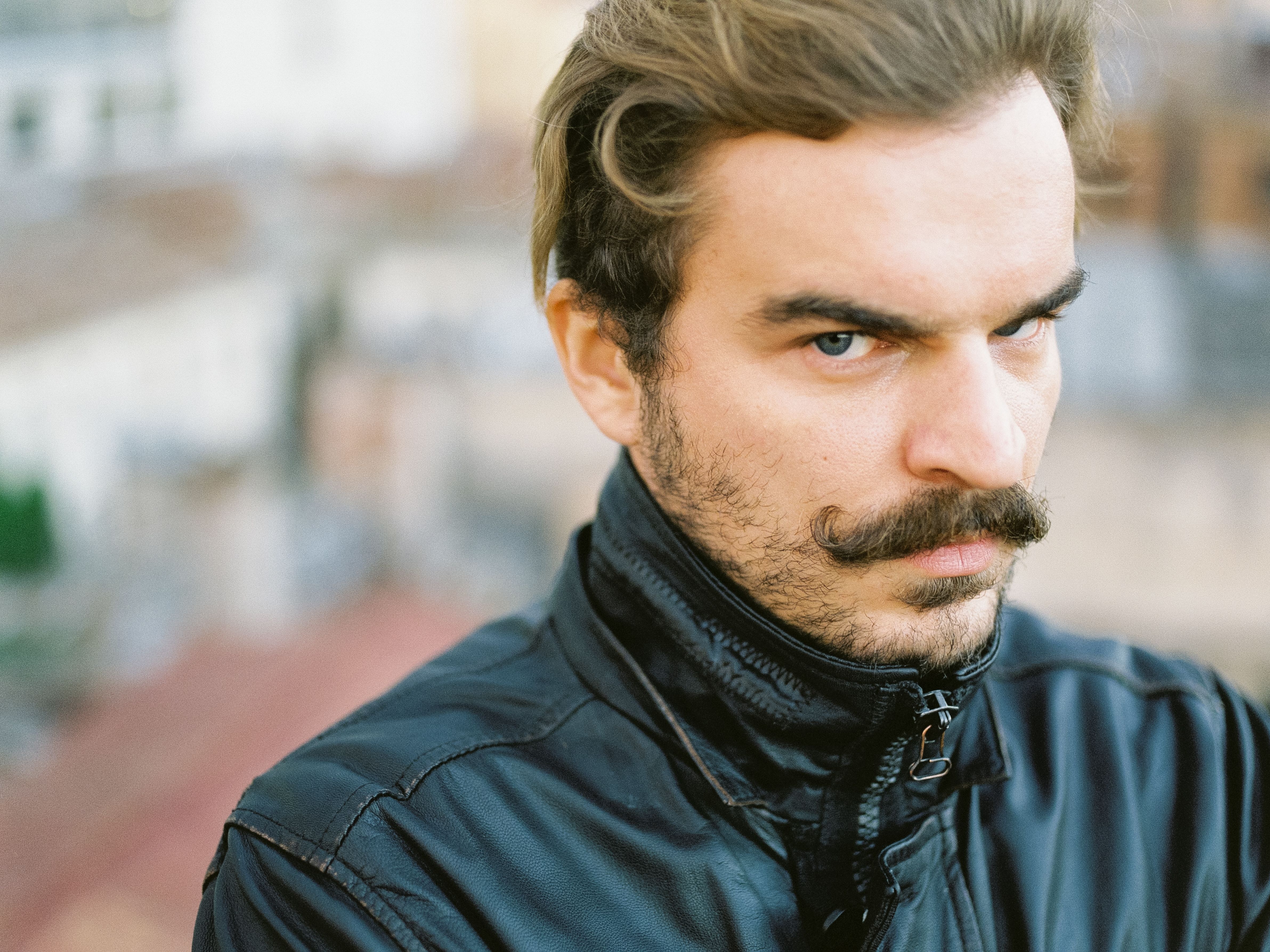 Close-up portrait of a man with a serious expression and mustache, wearing a jacket outdoors.