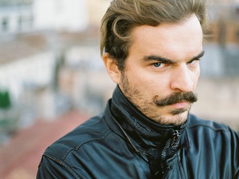 Close-up portrait of a man with a serious expression and mustache, wearing a jacket outdoors.
