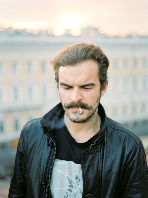 Free A man with a beard wearing a black leather jacket smokes a cigarette outdoors with a blurred cityscape background. Stock Photo