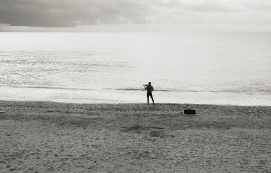 Grayscale photo of a lone fisherman casting his line on a serene beach.