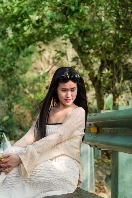An Asian woman sits by a guard rail in a forest, looking contemplative and relaxed.