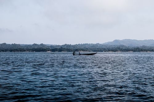 Free A small boat glides across a tranquil lake with distant forested hills under a cloudy sky. Stock Photo