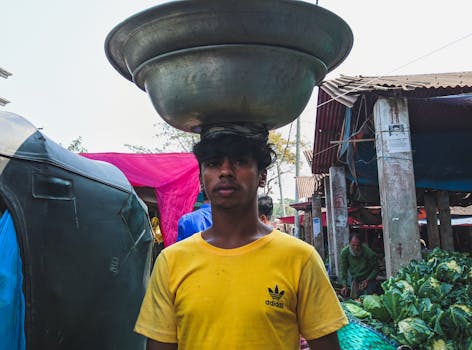 Young man balances a metal basin on his head at a Bangladesh market, showcasing traditional skills.