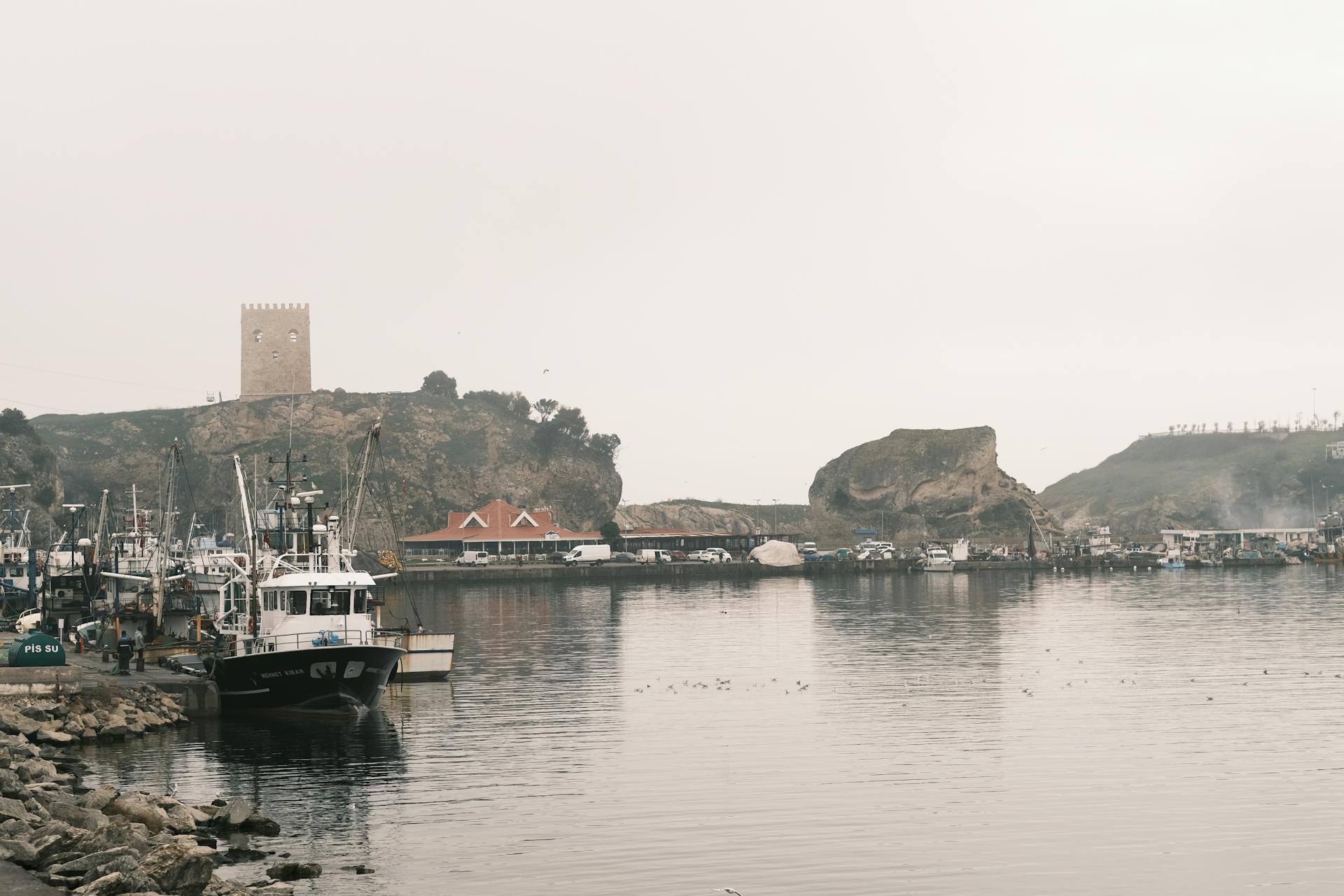 A tranquil misty harbor scene with moored boats and a stone tower in the distance.