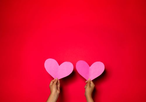 Free Two hands holding pink heart cutouts against a bold red backdrop, symbolizing love and romance. Stock Photo