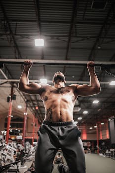 Shirtless bodybuilder showcasing strength during an intense workout on a pull-up bar in a gym.