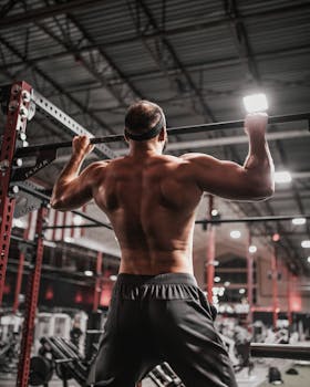Shirtless man doing pull-ups in a well-equipped gym, showcasing strength and fitness.