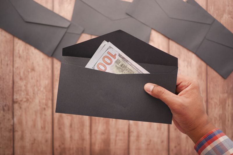 A hand holds a black envelope containing a 100 dollar bill on a wooden surface.