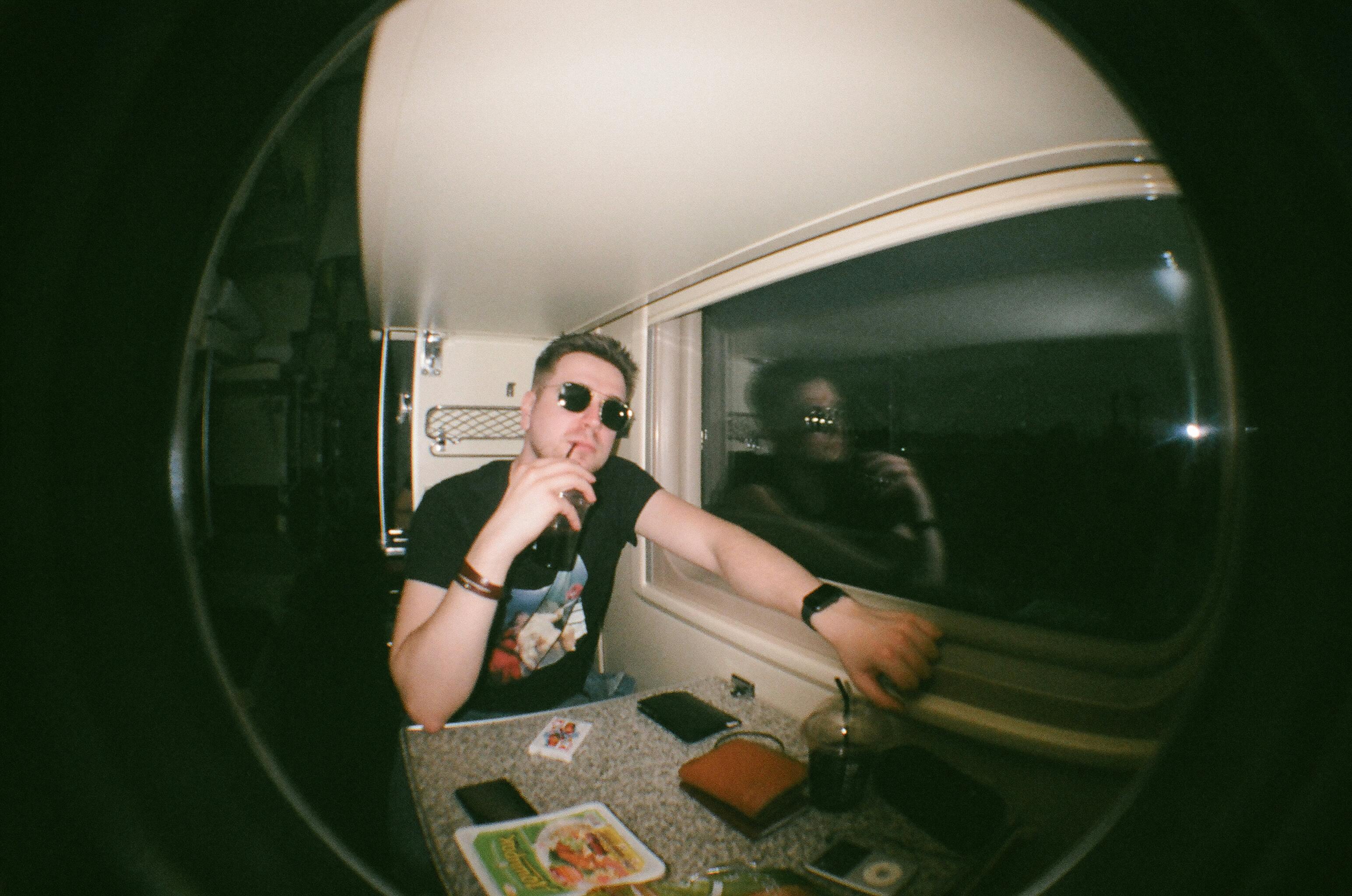 Man in sunglasses ponders by train window at night with reflection visible in glass.