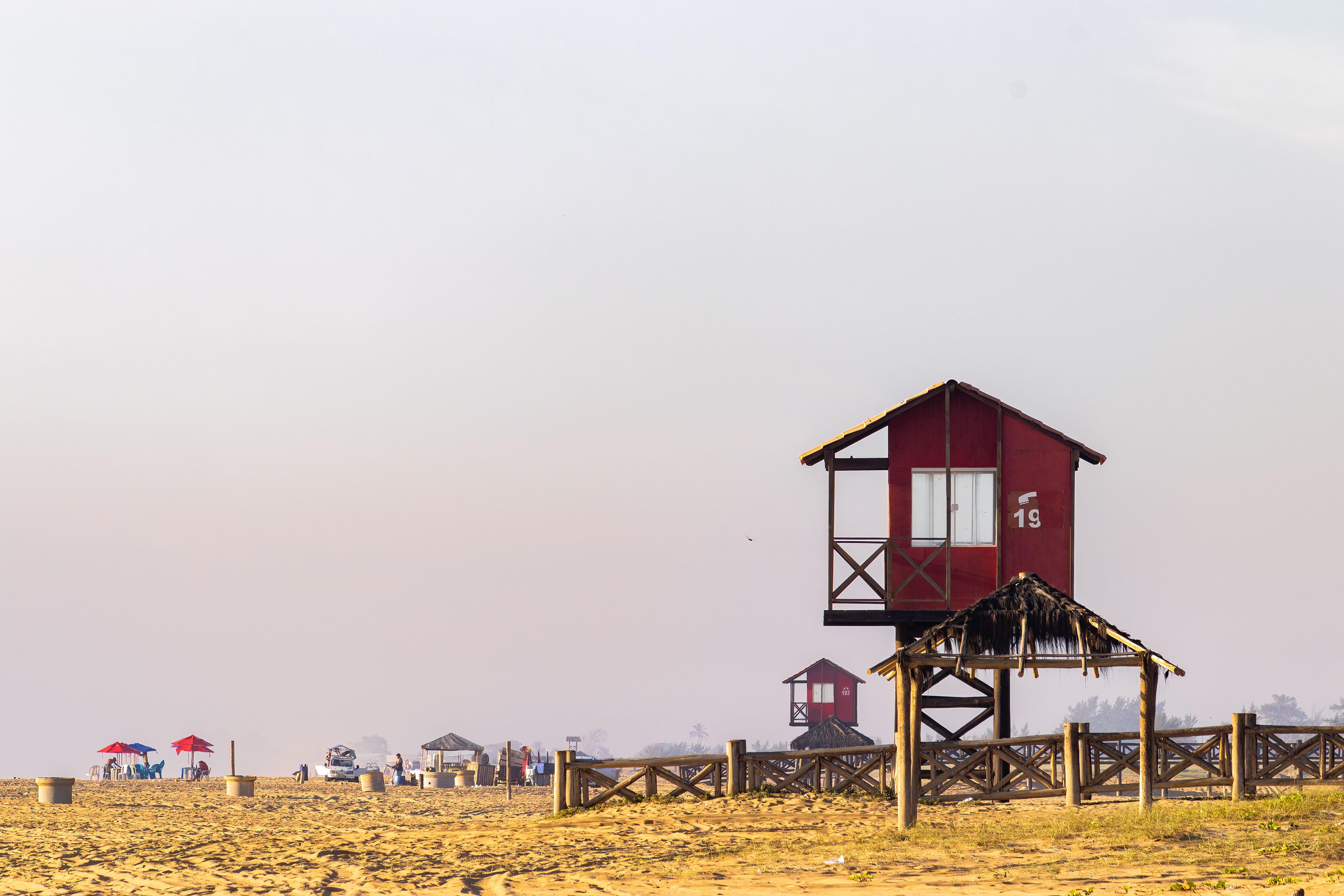 Red lifeguard tower on a sunny Brazilian beach with white sky and umbrellas in view.