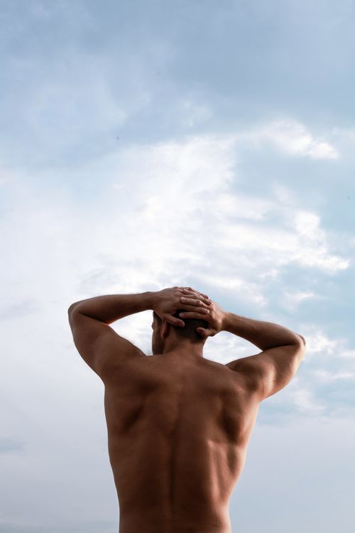 Free Back view of a muscular shirtless man with hands on head, set against a clear blue sky. Stock Photo