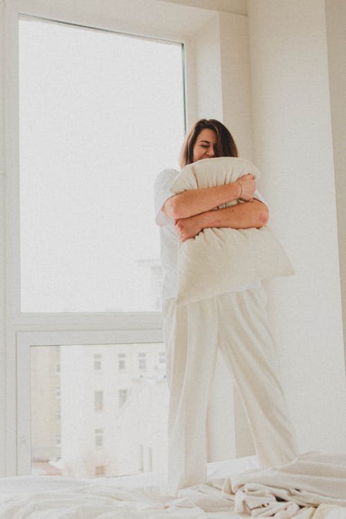 Woman in White Sleepwear Hugging a Pillow while Standing on the Bed