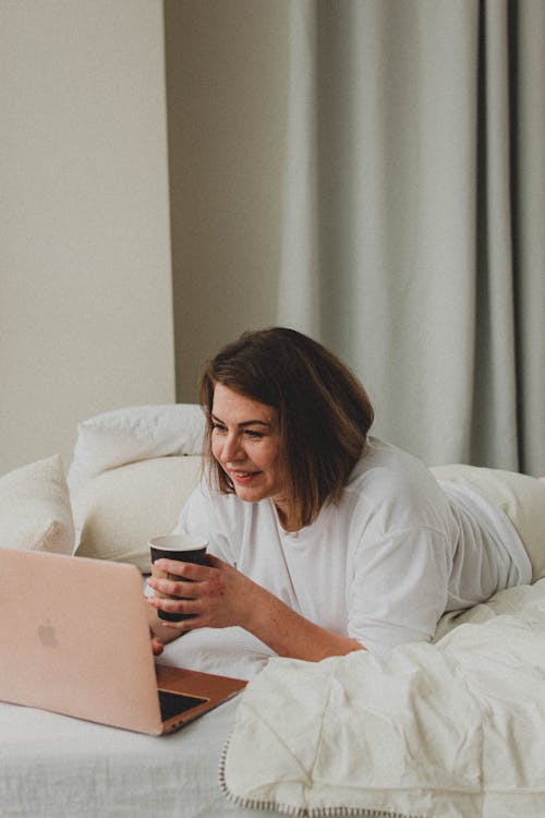 Woman Lying in Bed with Laptop and Cup of Coffee