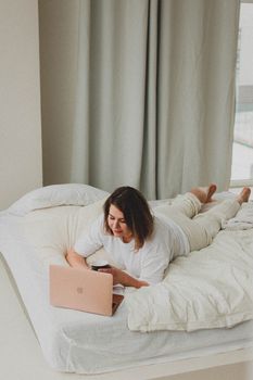 Woman lying on bed with laptop, comfortably working from home, enjoying a coffee break.