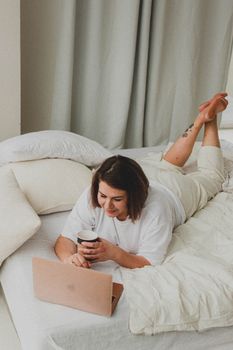 Woman lying on a bed with a laptop and coffee, enjoying a cozy morning indoors.