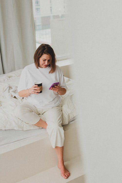 Woman Sitting in White Shirt Holding Cup of Coffee and Using Smartphone