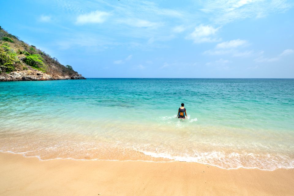 Person wading into calm turquoise water from a sandy beach cove