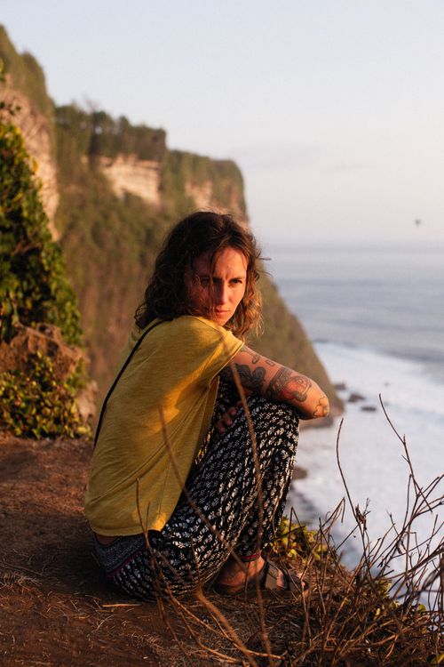 Free A tattooed woman sitting on a cliff edge overlooking the ocean during sunset. Stock Photo