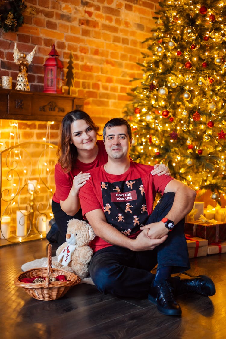 A Couple In Red Shirts Sitting Beside A Christmas Tree