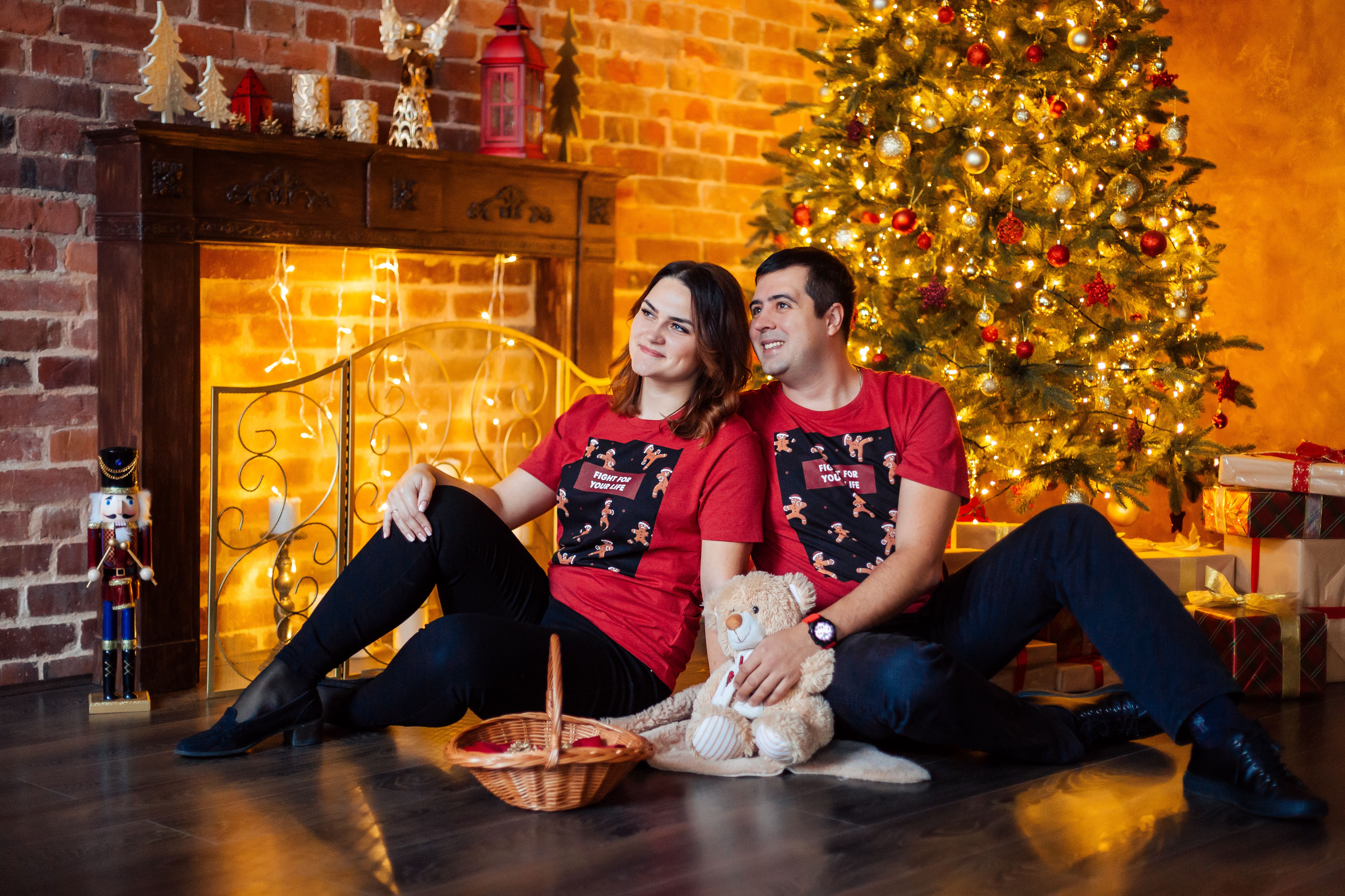 A smiling couple in matching red shirts enjoys a cozy Christmas moment by a glowing tree indoors.