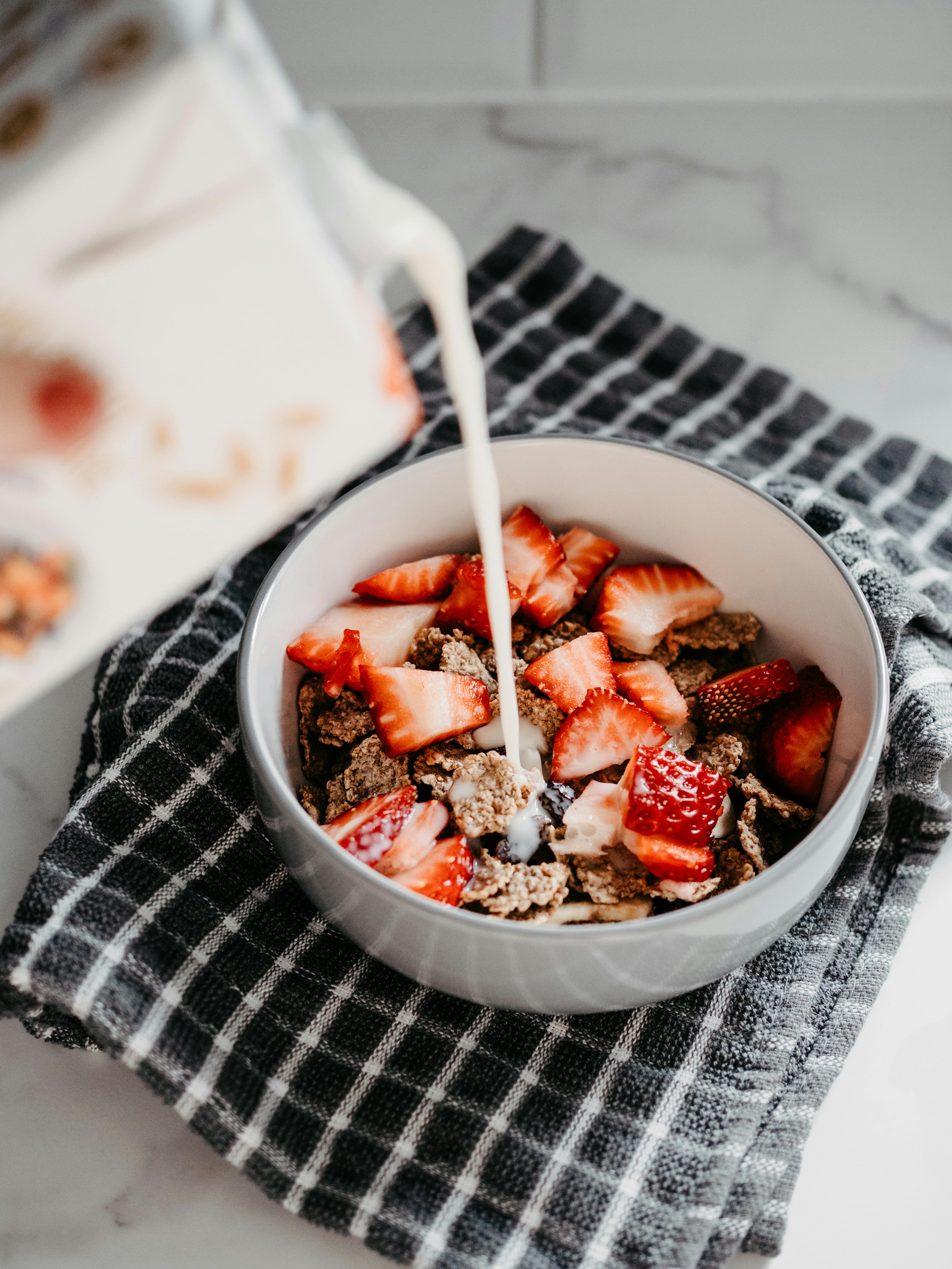 Close-up of milk being poured over cereal with fresh strawberries on a checkered cloth indoors.