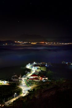 A mesmerizing aerial view of a village illuminated at night, surrounded by mountains and soft fog.