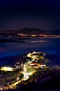 Illuminated mountain village at night with fog and distant city lights.