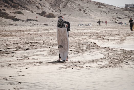Surfer with a surfboard on the sandy shores of El Cotillo, Spain, surrounded by ocean and dunes.