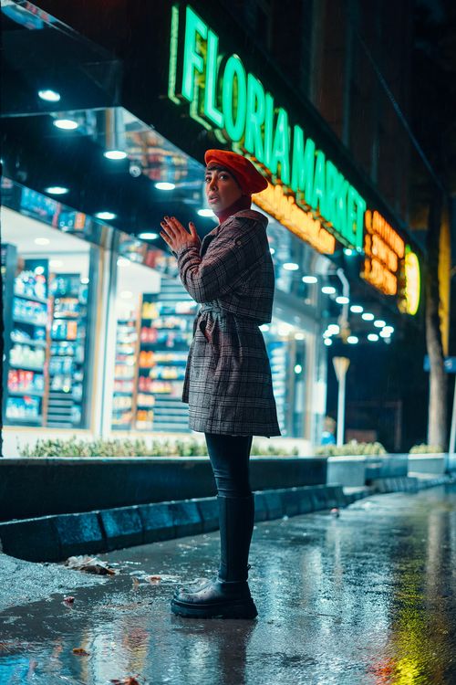 Free Stylishly dressed woman in a beret stands by a neon-lit Parisian market during a rainy night. Stock Photo
