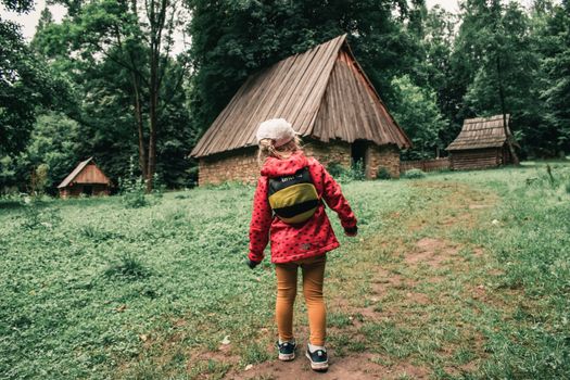 A young girl walks toward wooden houses in a lush green setting, Sanok, Poland.