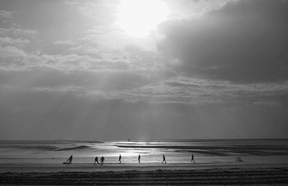 Silhouettes play soccer on La Baule beach under a dramatic cloudy sky at sunset.