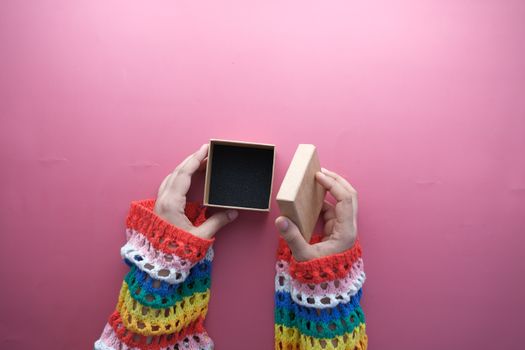 A close-up of hands in colorful sleeves opening a jewelry box on a pink background.