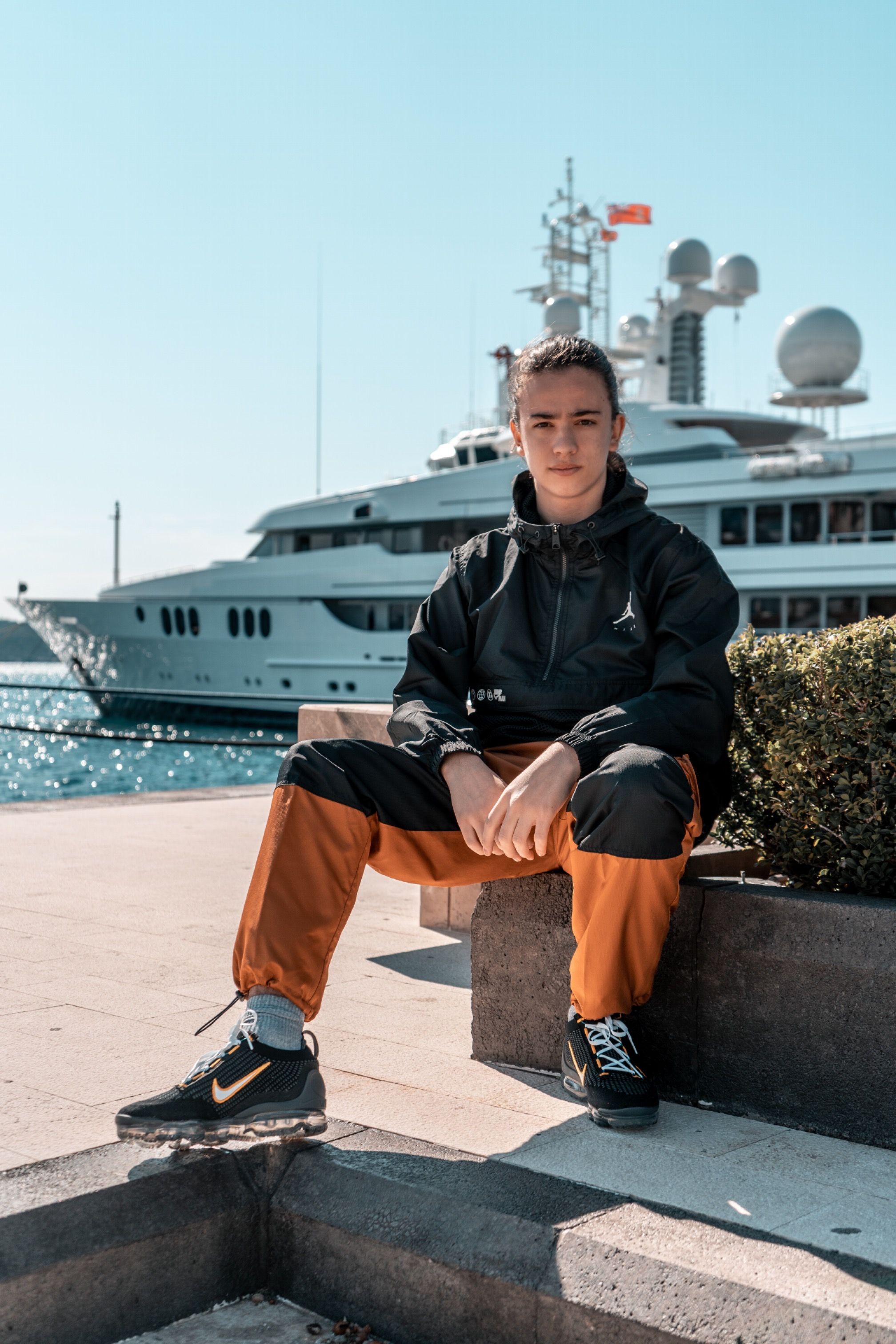 Young adult in trendy outfit sitting by a luxury yacht in Tivat, Montenegro.